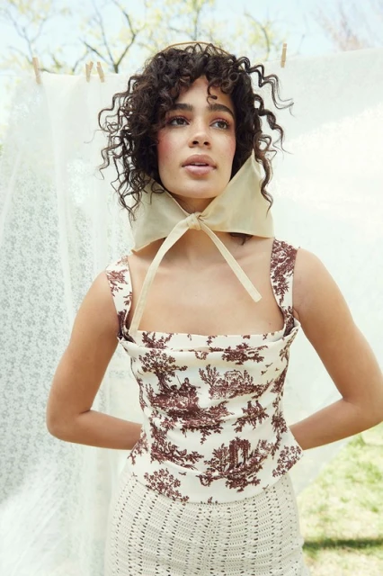 A woman poses outdoors wearing a patterned top and skirt, with a sheer scarf tied around her head, in front of a draped white sheet.