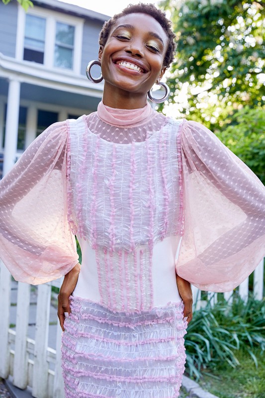 Smiling African American woman with hoop earrings stands outdoors in a sheer pink dress in front of a light blue house and green foliage.