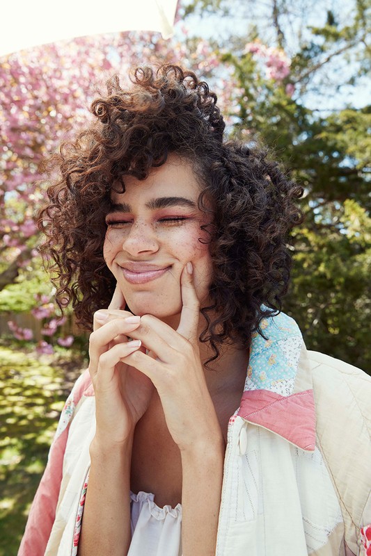 Close-up portrait of a smiling adult female with curly dark hair, her hands on her cheeks, wearing a patterned jacket in a natural outdoor setting.