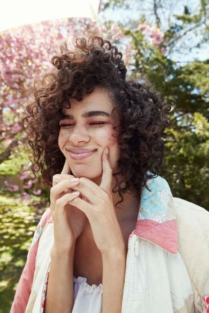 Close-up portrait of a smiling adult female with curly dark hair, her hands on her cheeks, wearing a patterned jacket in a natural outdoor setting.