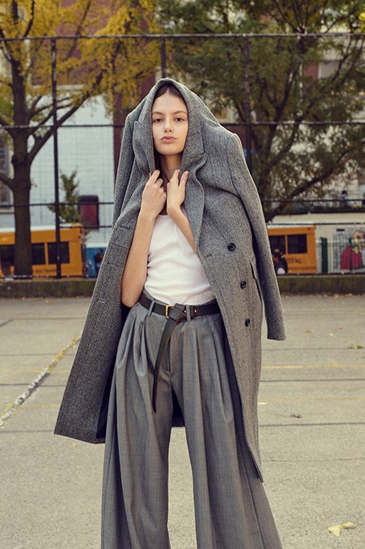 A woman poses in a white tank top, gray pants, and an oversized gray coat in an outdoor urban environment on a sunny day.