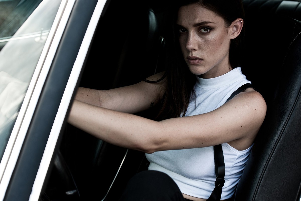 A woman with dark hair and a white top sits in a vintage car with a black interior, looking intently at the camera with a serious expression.