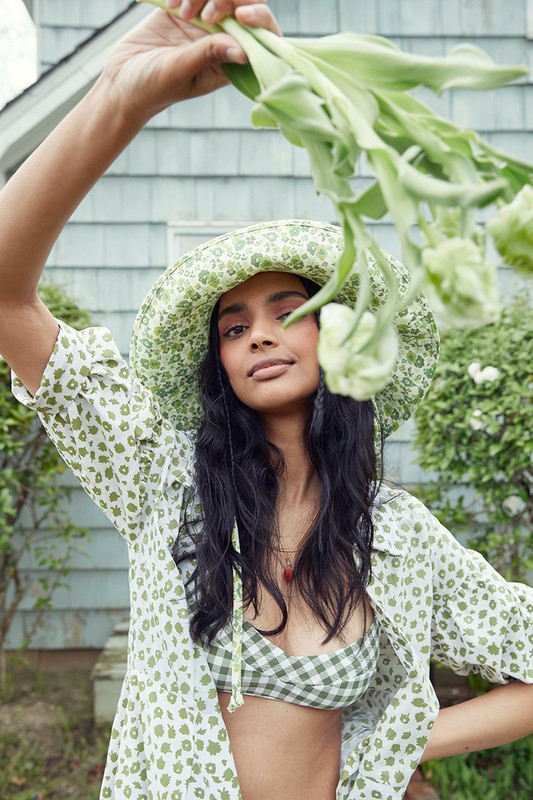 Woman in floral hat and bikini top holds flowers in outdoor setting, featuring summer fashion, natural light, and a cheerful mood.
