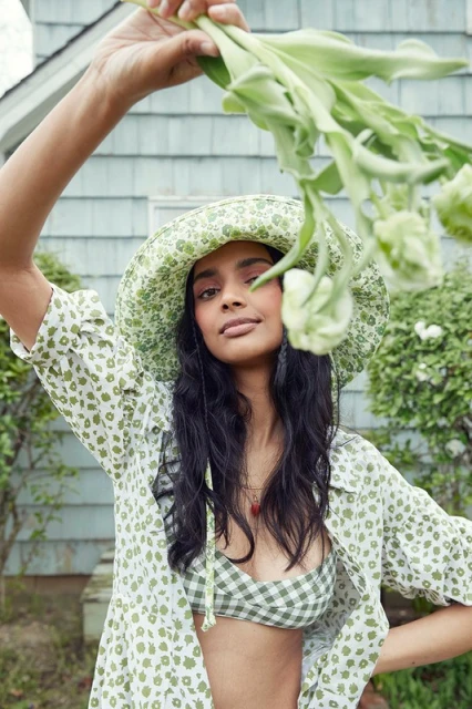 Woman in floral hat and bikini top holds flowers in outdoor setting, featuring summer fashion, natural light, and a cheerful mood.