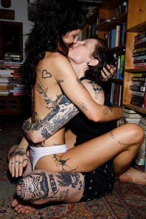 Two tattooed people embracing and kissing intimately near a wooden bookcase inside a room styled by wardrobe stylist Morgan Gibbons.