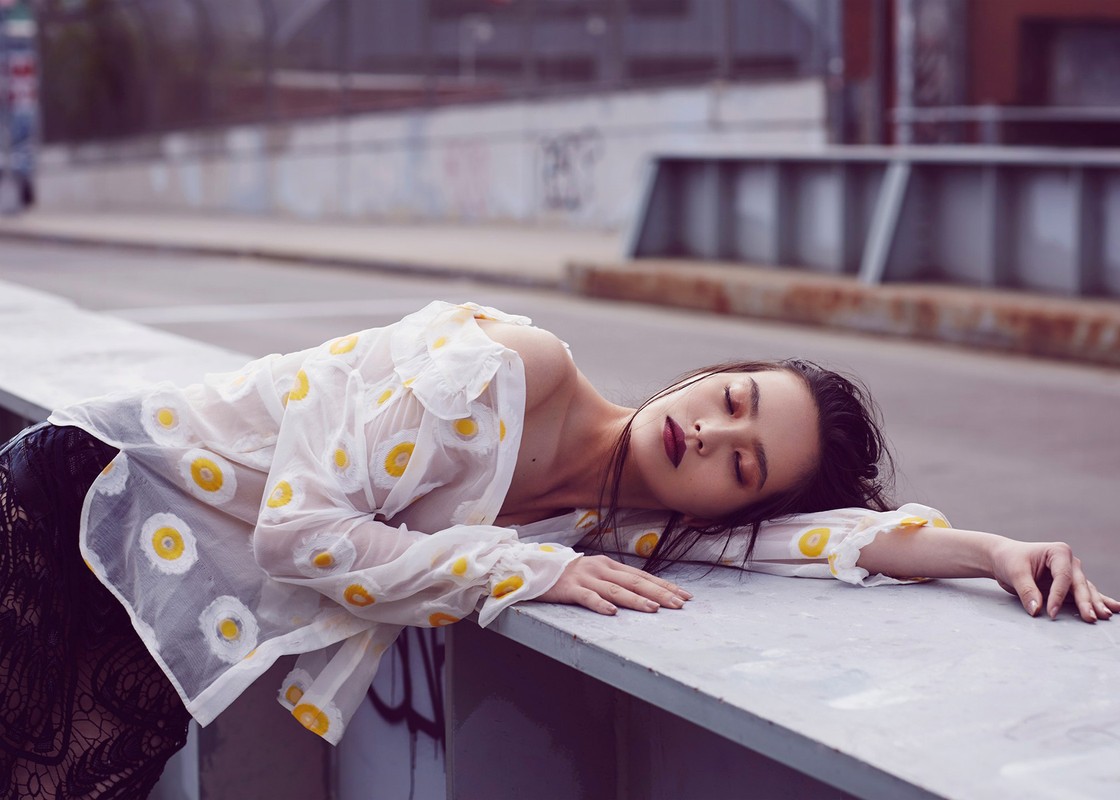 Adult female model poses in a sheer white daisy-print shirt and dark lace skirt against an urban bridge backdrop with soft, diffused lighting.