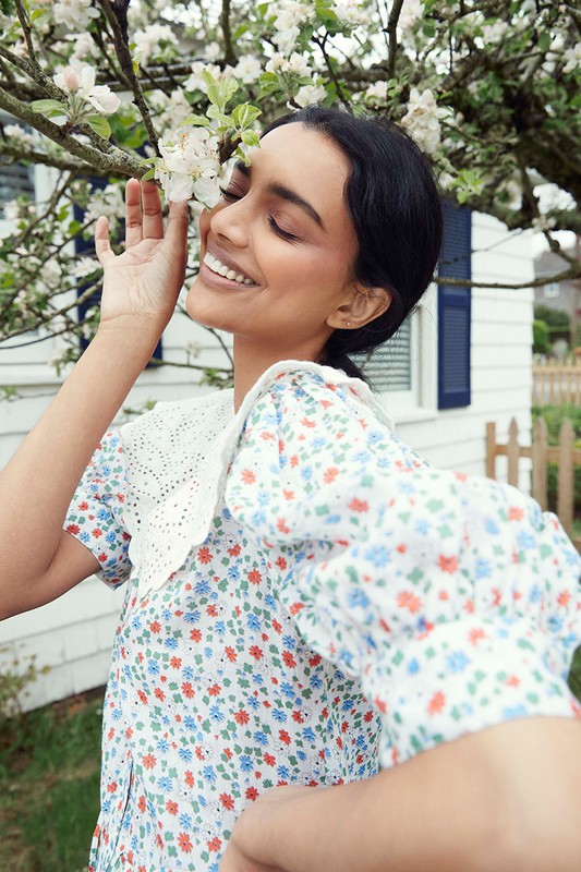 Smiling woman in a floral dress touches white blossoms on a tree branch outside of a white house with blue shutters on a sunny day.