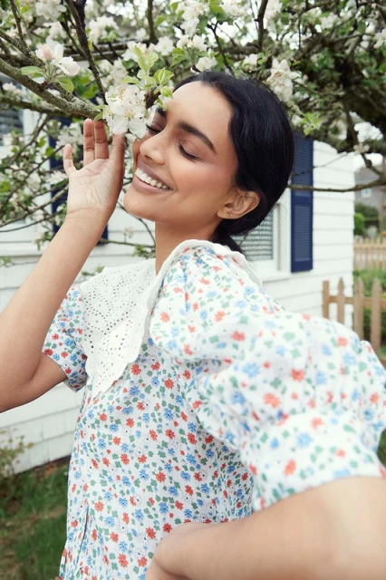 Smiling woman in a floral dress touches white blossoms on a tree branch outside of a white house with blue shutters on a sunny day.