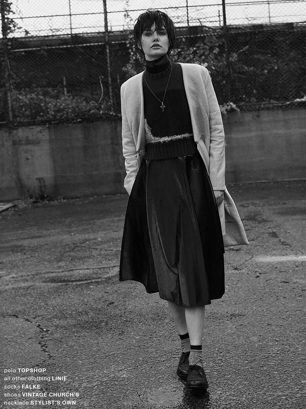 A woman in a black dress, socks, and shoes stands on a city street, wearing a beige coat and silver necklace, in a monochromatic fashion shot.