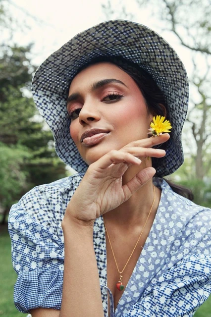Portrait of a South Asian adult woman in a patterned bucket hat, wearing a floral print dress, with a yellow flower by her ear in a green outdoor setting.