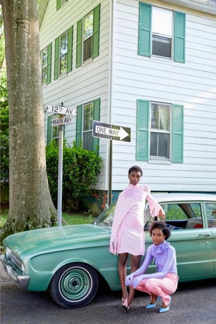 Two women pose with a vintage car on a street with a house in the background. The women wear colorful outfits in an outdoor setting.