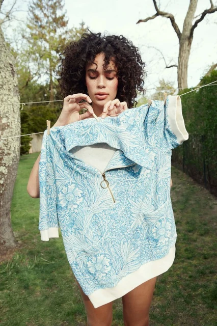 Brunette woman hangs a blue floral-patterned hoodie on a clothesline outdoors on a sunny day, surrounded by green grass and trees.