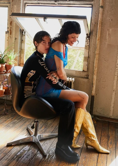 Two women in an embrace on a black chair. The blonde is wearing a blue dress and gold boots, while the other is in black pants and shirt.