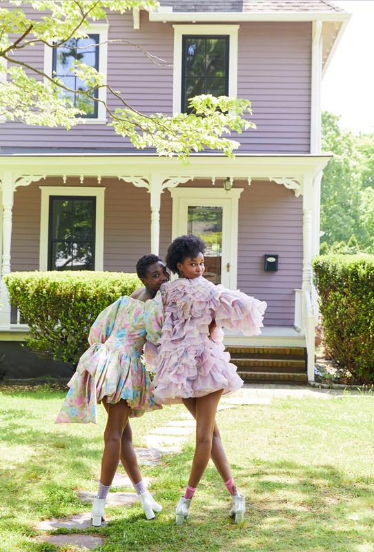Two young Black women pose in colorful frilly dresses on a lawn in front of a lilac house, showcasing fashion and style in an outdoor setting.