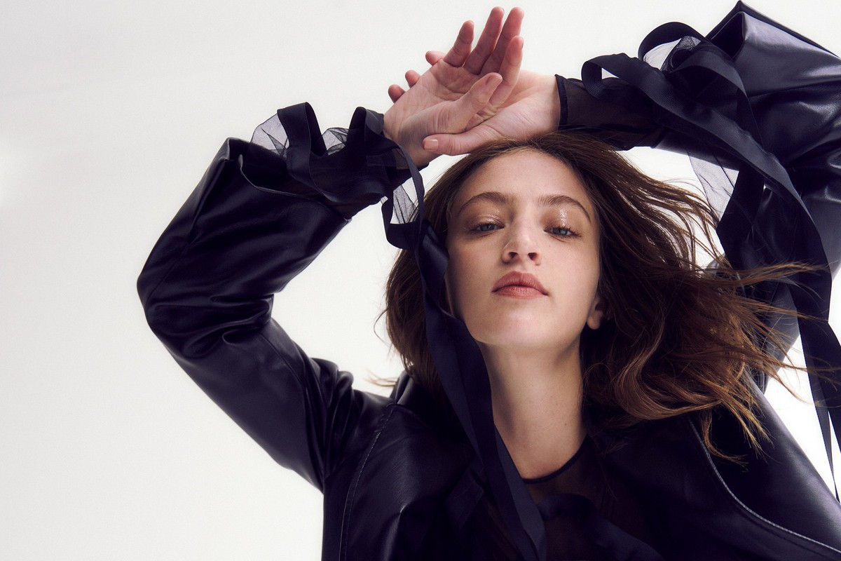 Fashion shot of a woman wearing a black jacket in a studio. The portrait features an upward angle and a bright neutral background.