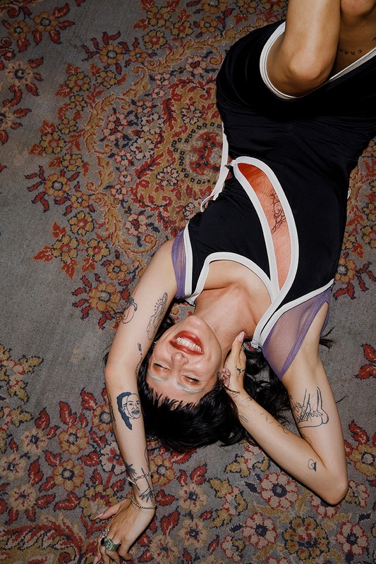 Woman with tattoos in fashion clothing lies on patterned carpet, smiling, indoors. Overhead angle captures bold colours, vintage look.