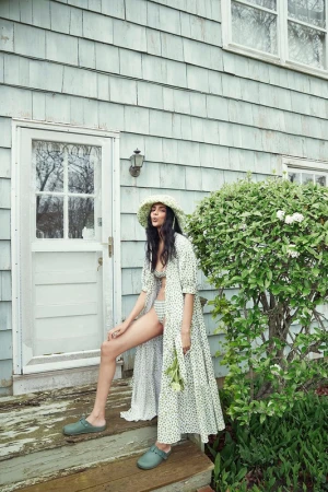 A woman in a dotted robe and sun hat poses on wooden steps outside a blue clapboard house next to a green bush in the daytime.