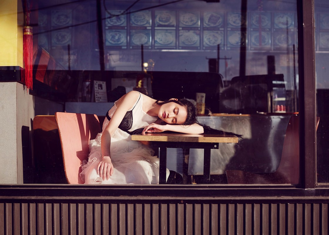 Young adult woman with long dark hair rests head on table inside restaurant booth with a dress; reflections on the window.