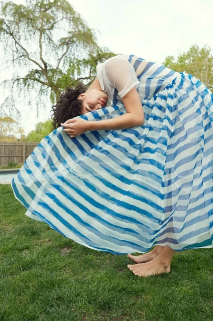 Woman in a blue and white striped dress poses barefoot in a backyard with grass and trees, creating a whimsical fashion image.