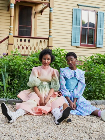 Two Black women in vintage dresses pose in front of a yellow house with light blue shutters, seated on the gravel ground with green plants.