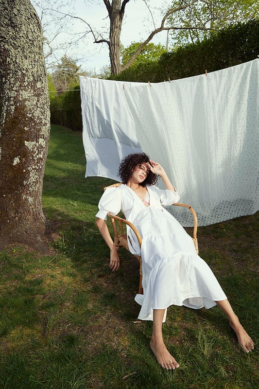 Woman with curly dark hair wearing a white dress seated in a wooden chair on grass outdoors with a laundry line behind her.