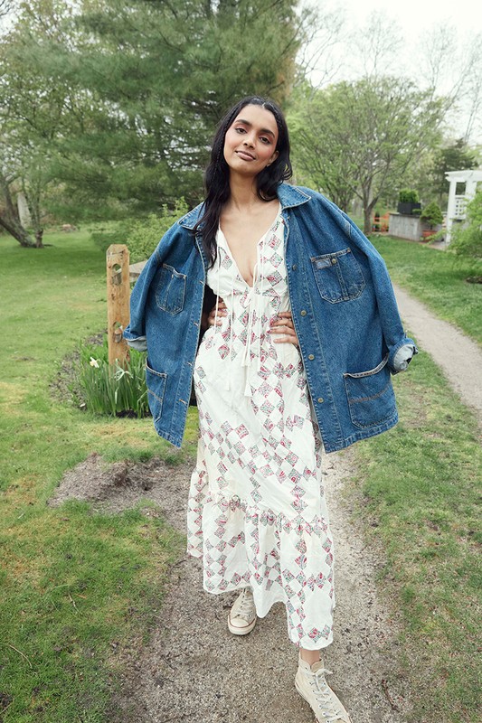 A full shot of a woman with her hands on her hips, wearing a white patterned dress and jean jacket, standing in a grassy outdoor setting.