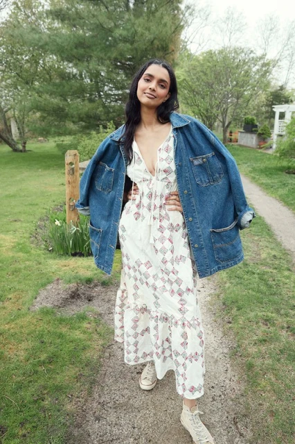 A full shot of a woman with her hands on her hips, wearing a white patterned dress and jean jacket, standing in a grassy outdoor setting.