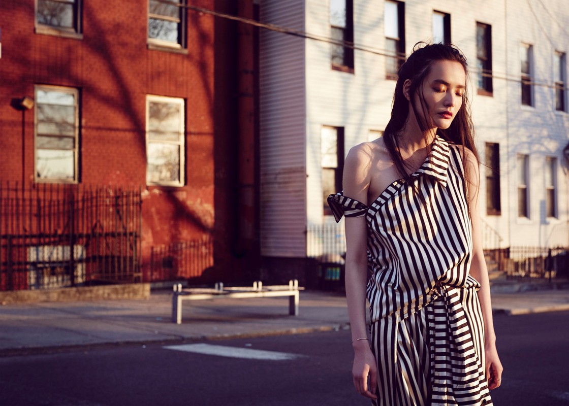 Asian woman stands on urban street in black and white striped off-shoulder dress with brick buildings in the background in sunlight.