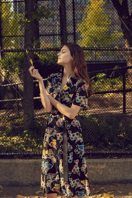 Adult female in floral dress stands outdoors near trees and chain link fence holding a small branch in natural light.