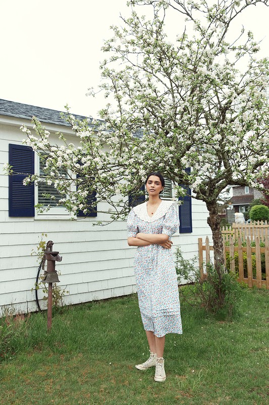 Adult female in a floral dress stands in a grassy yard next to a blooming tree and a white house with dark shutters.