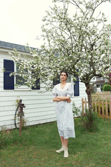 Adult female in a floral dress stands in a grassy yard next to a blooming tree and a white house with dark shutters.