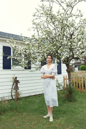 Adult female in a floral dress stands in a grassy yard next to a blooming tree and a white house with dark shutters.