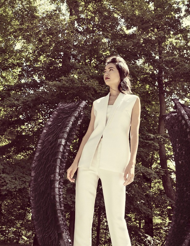 Stylish Asian woman in an off-white pantsuit poses outdoors next to a sculpture in a wooded area on a sunny day.