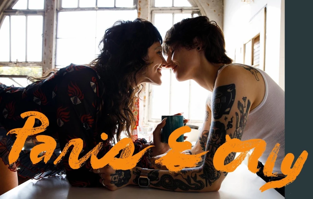 Two adult women, close together indoors, one holding a mug, with windows providing natural light. The women are smiling and affectionate.