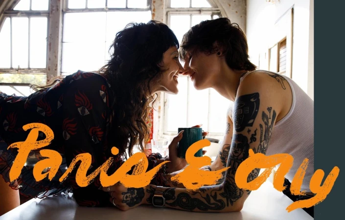 Two people with tattoos leaning in close to touch noses indoors with large bright windows in the background.
