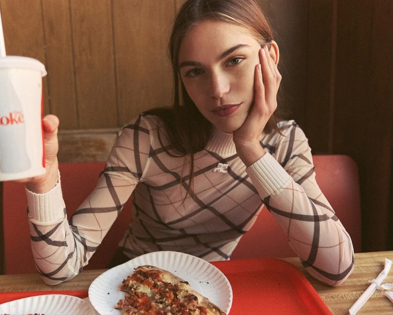 Young woman in a plaid top sitting at a booth in a diner, eating pizza and holding a cup with a straw. Indoor setting.