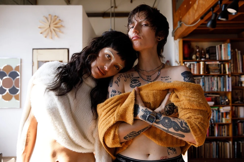 Two tattooed people pose indoors; a woman rests her head on the shoulder of a person with an orange sweater and Calvin Klein waistband.