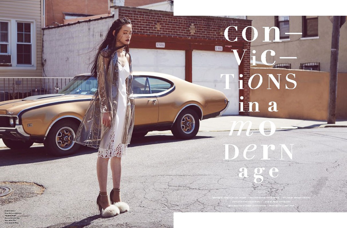 Adult female poses on urban street next to vintage gold car, wearing transparent raincoat and white dress, medium shot in daylight.