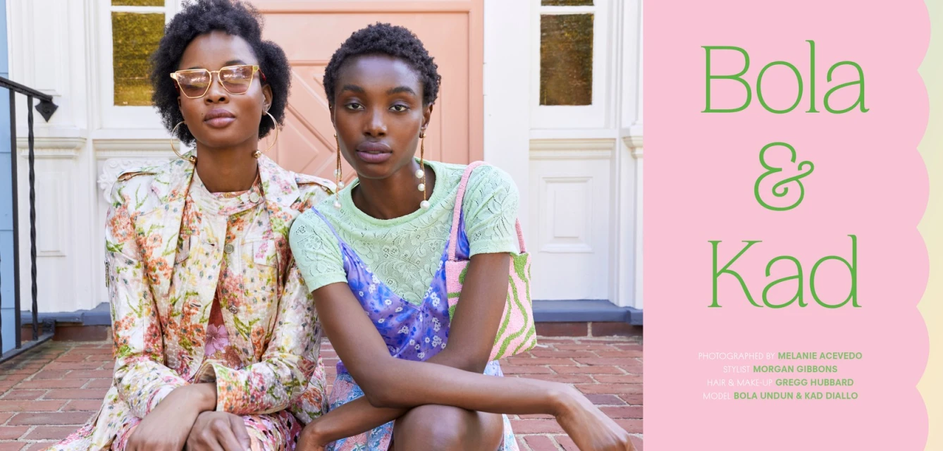 Two African-American women pose on porch steps, adorned in floral patterns, stylish dresses, and sunglasses in soft daylight.