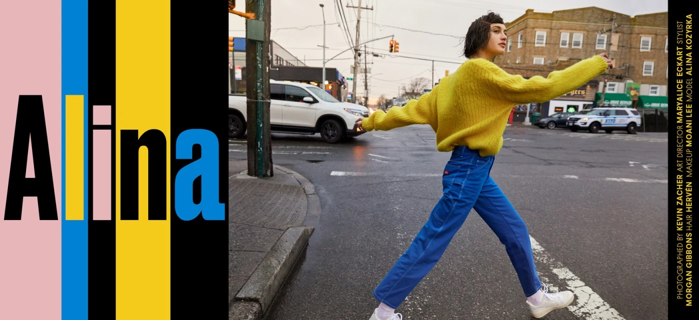 A young woman in a yellow sweater and blue pants strides across a city crosswalk, cars in the background, with vertical color blocks on the left.