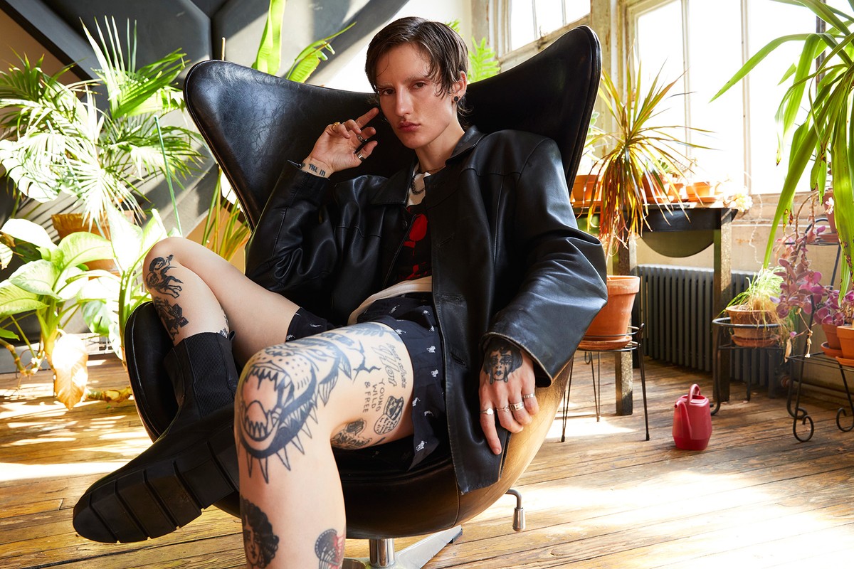 An adult with tattoos relaxes in an egg chair indoors surrounded by potted plants, wearing a black leather jacket and shorts.