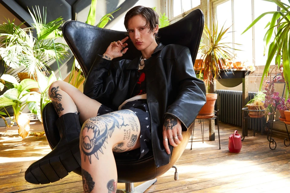 An adult with tattoos relaxes in an egg chair indoors surrounded by potted plants, wearing a black leather jacket and shorts.