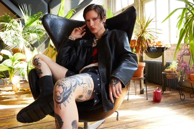 An adult with tattoos relaxes in an egg chair indoors surrounded by potted plants, wearing a black leather jacket and shorts.