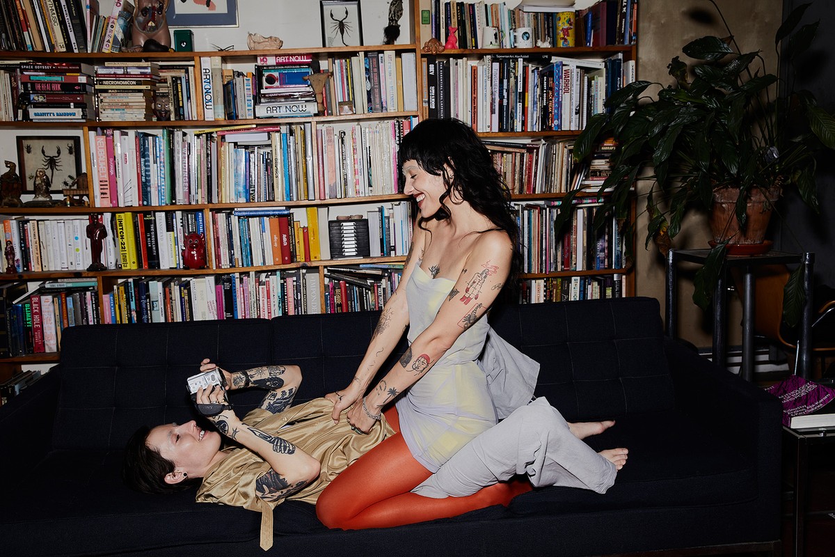 Two tattooed women on a couch, one lying down taking a photo, the other sitting, laughing, wearing grey and orange leggings. 