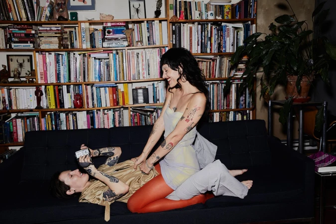 Two tattooed women on a couch, one lying down taking a photo, the other sitting, laughing, wearing grey and orange leggings. 