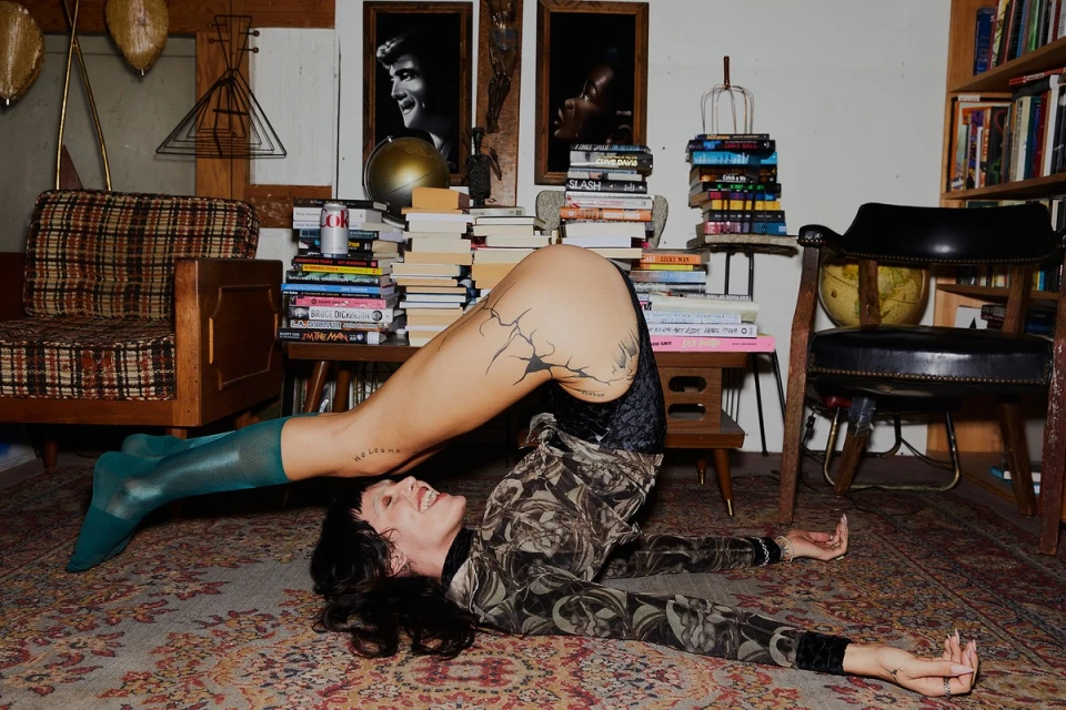 Adult woman in artistic yoga pose on patterned rug, surrounded by piles of books, vintage furniture, and framed portrait art.
