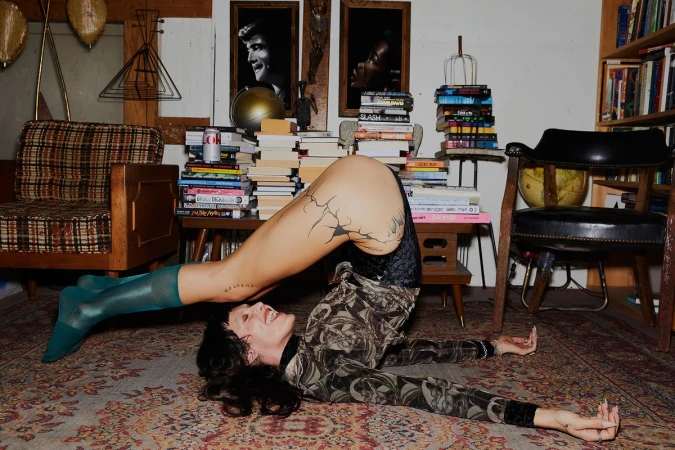 Adult woman in artistic yoga pose on patterned rug, surrounded by piles of books, vintage furniture, and framed portrait art.
