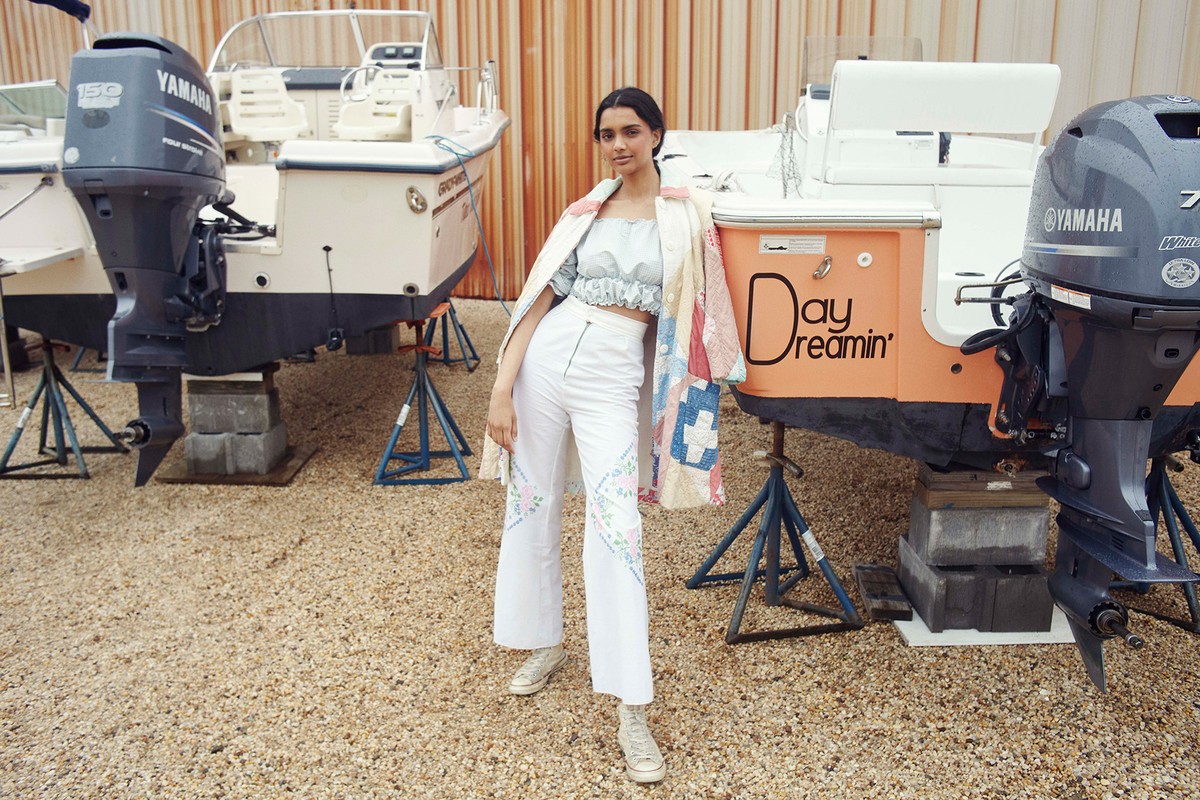 Woman in embroidered pants poses on a gravel surface in front of boats supported by stands, wearing a patchwork-style jacket.