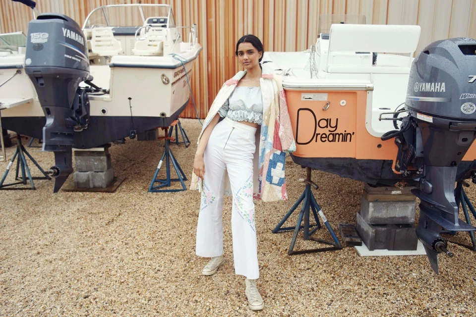 Woman in embroidered pants poses on a gravel surface in front of boats supported by stands, wearing a patchwork-style jacket.