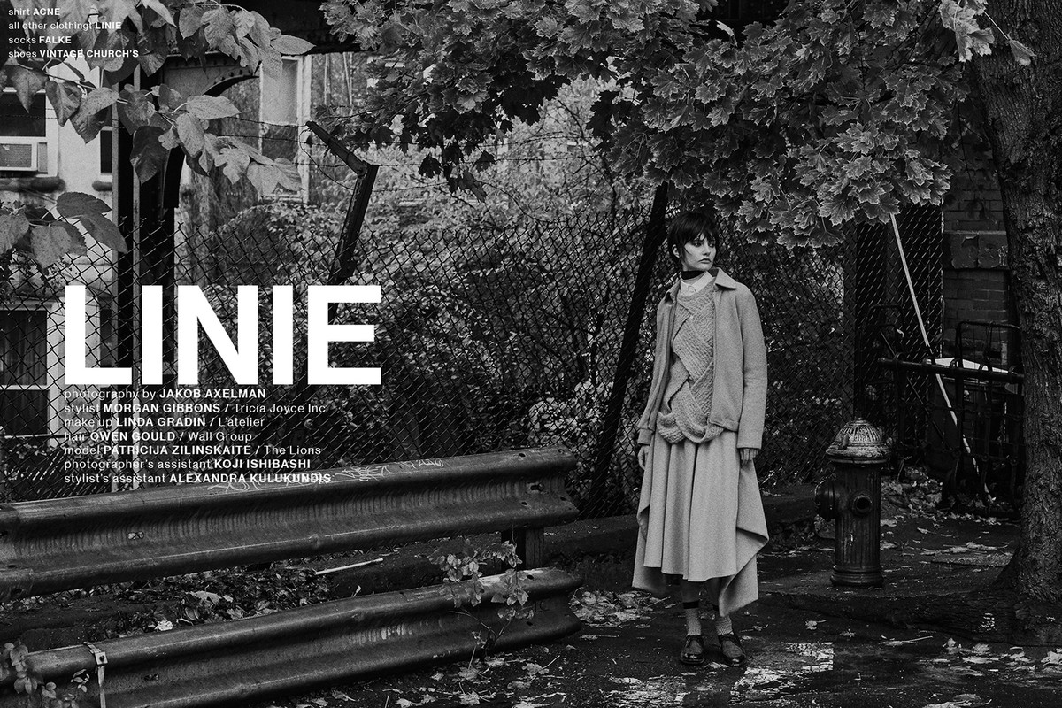 Black and white fashion shot of a woman in a coat and skirt standing outside with trees and a fence creating a natural yet urban backdrop.